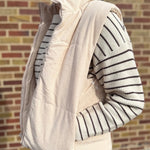 Person wearing a cream puffer vest over a striped sweater against a brick wall.