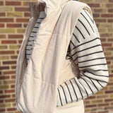 Person wearing a cream puffer vest over a striped sweater against a brick wall.