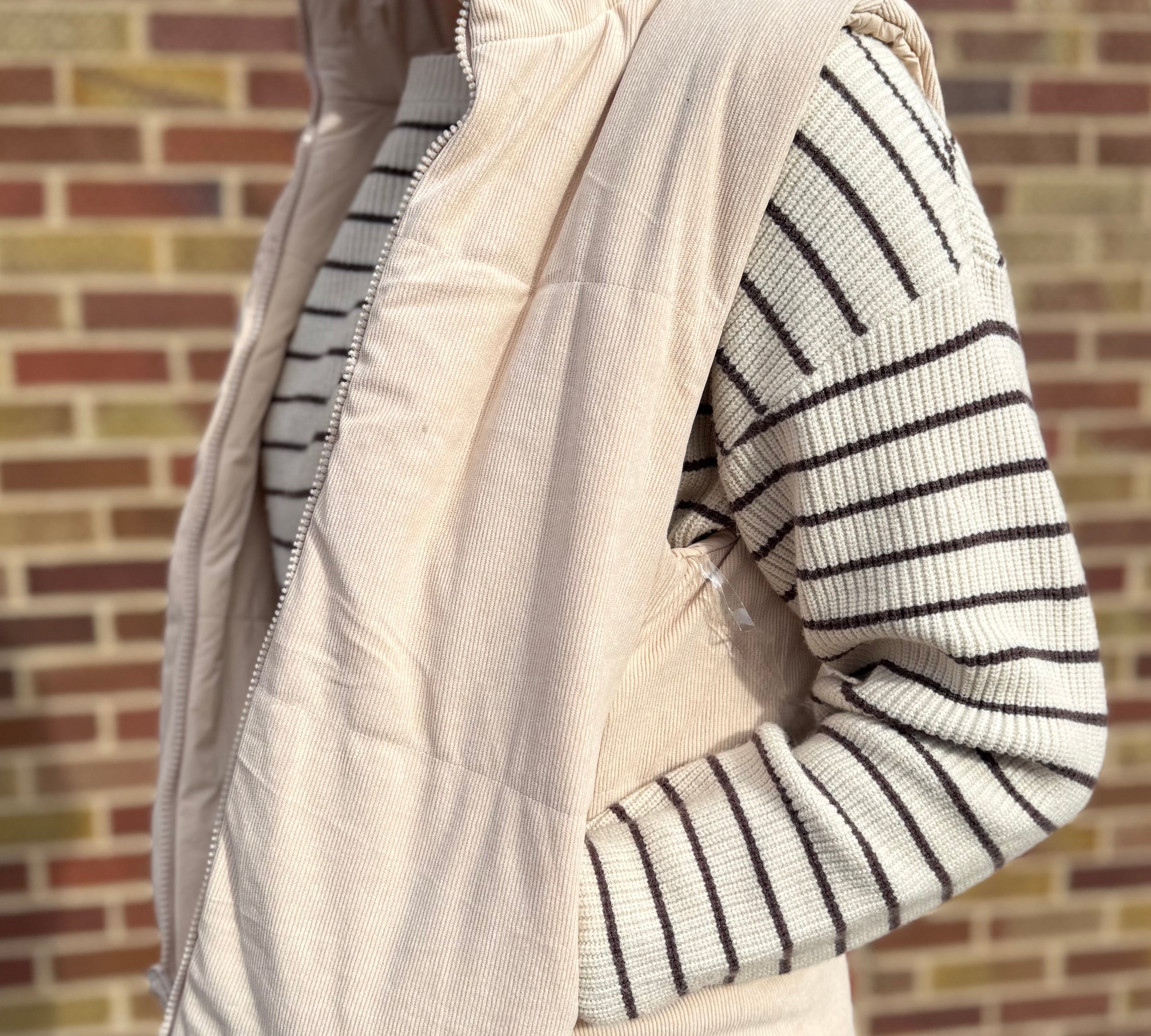 Person wearing a cream puffer vest over a striped sweater against a brick wall.