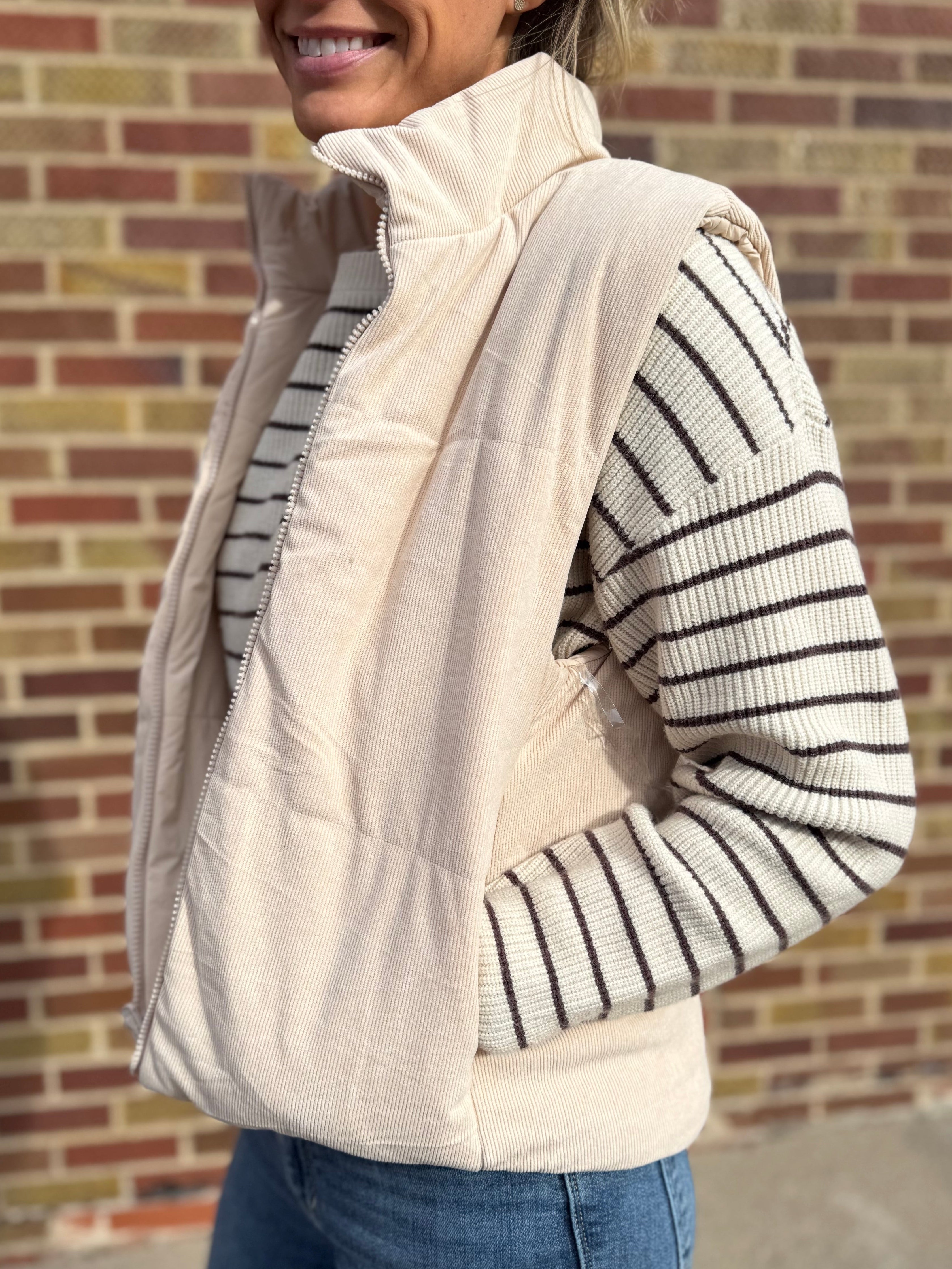 Person wearing a cream puffer vest over a striped sweater against a brick wall.